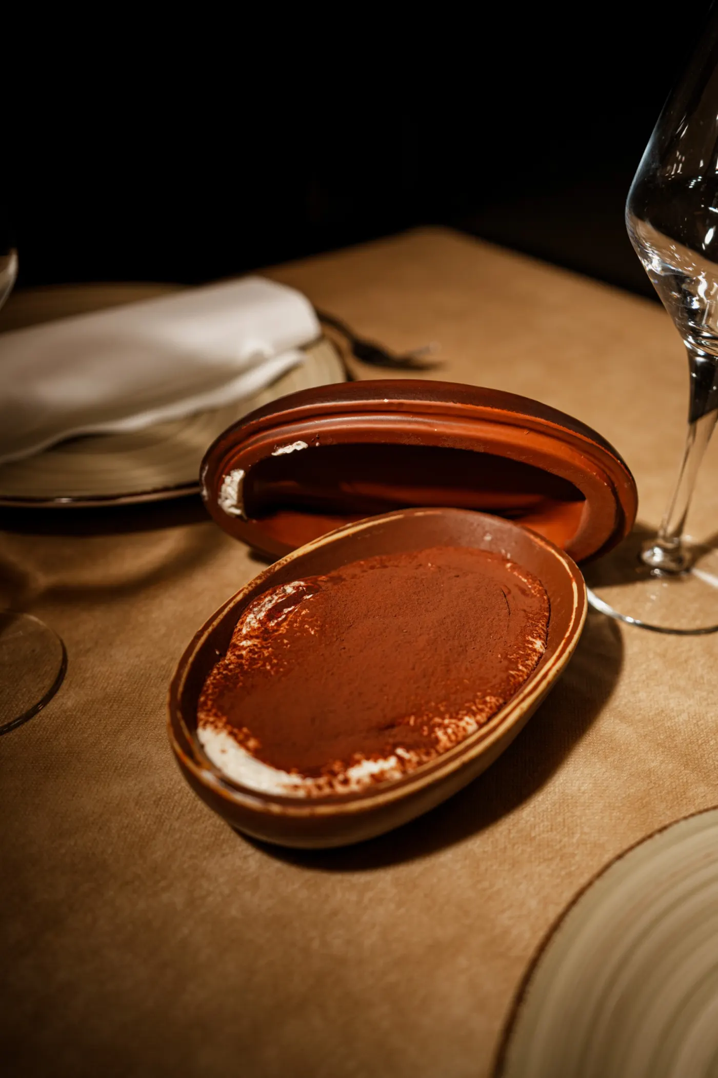 Tiramisú espolvoreado de cacao en una fuente ovalada de terracota, con la tapa abierta al lado, sobre una mesa puesta.